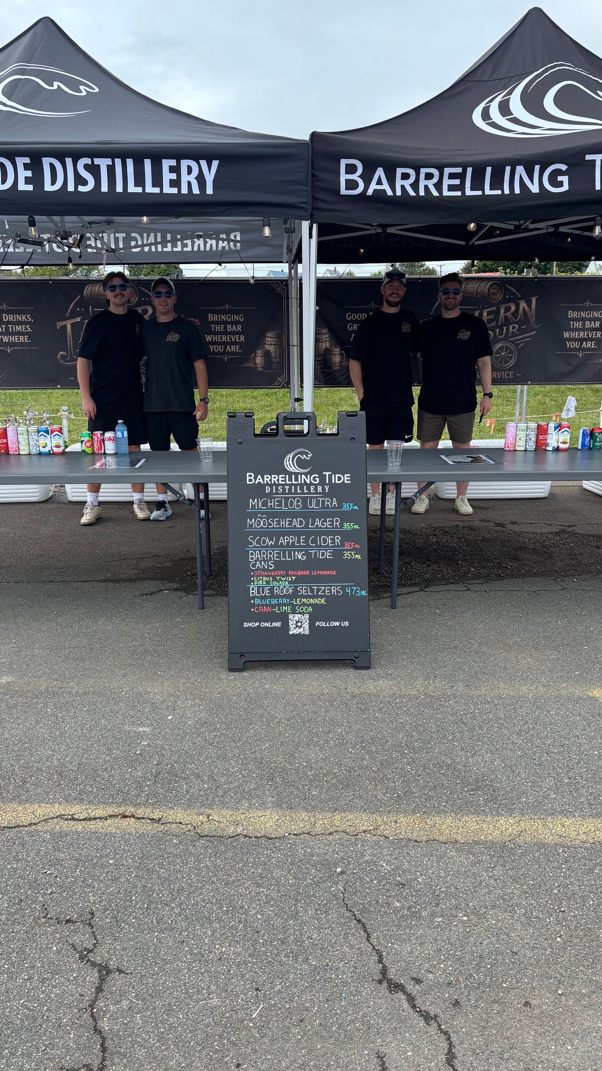 A Barrelling Tide Distillery outdoor booth setup on asphalt under a black canopy tent, with three people standing behind a table and a chalkboard sign in front.
