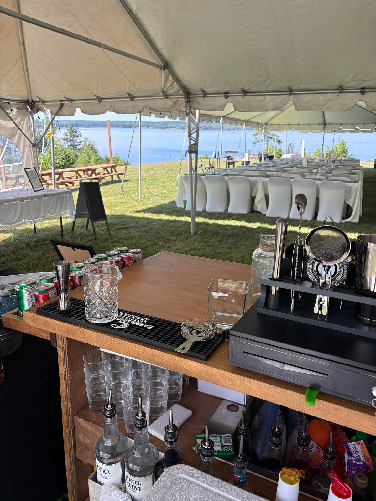 A lakeside outdoor bar setup under a large white event tent, with tables and chairs arranged in the background on the grass. The bar counter holds mixers, bottles, glasses, a strainer, and a black bar mat with white text. on the Mira river in cape breton