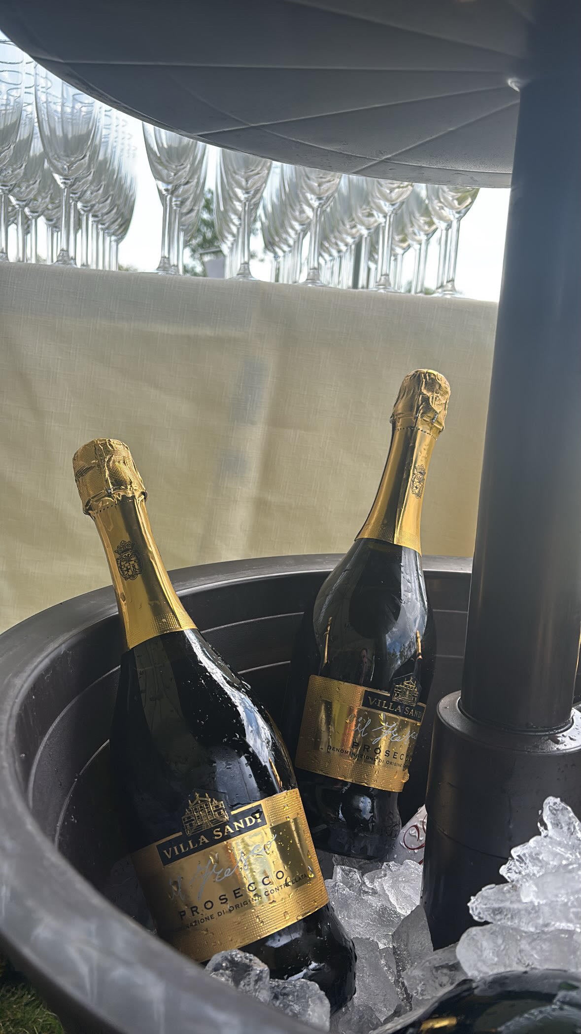 Two bottles of Villa Sando Prosecco resting in a black ice bucket filled with crushed ice, with a blurred background of hanging wine glasses and a cream tablecloth.