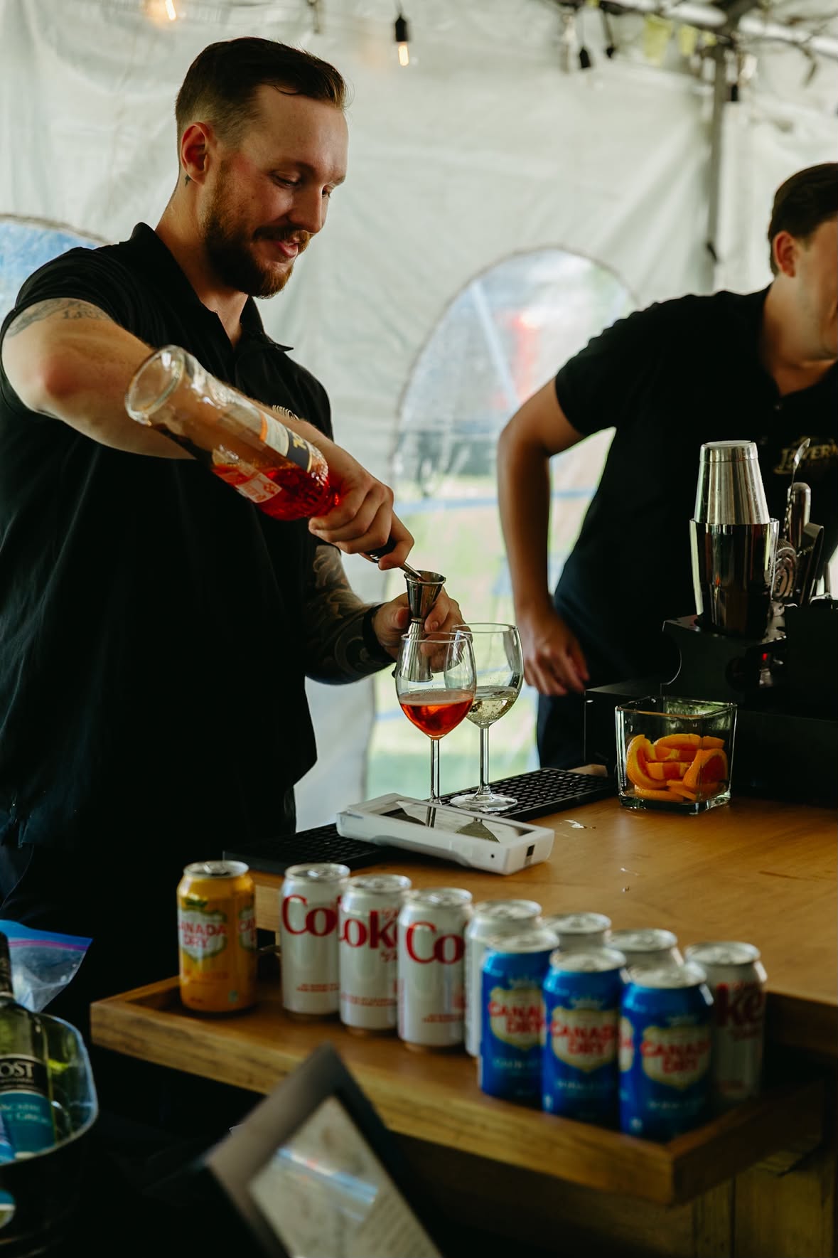 A bartender in a black shirt pours a pink drink from a bottle into two wine glasses on a wooden bar counter, with various canned sodas and a cocktail shaker nearby in a tent setting.